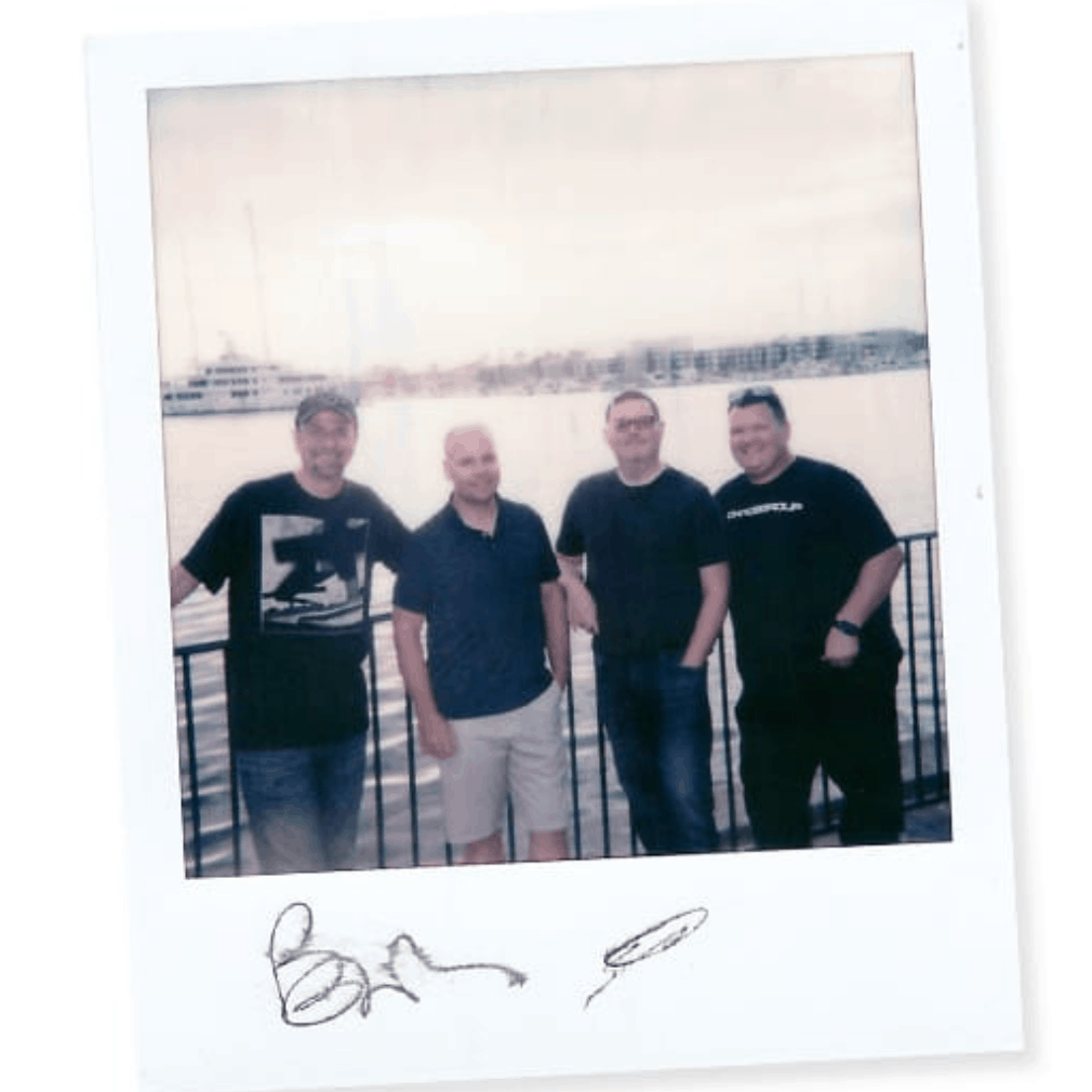 Four casually dressed men posing by waterfront railing with marina in soft daylight background