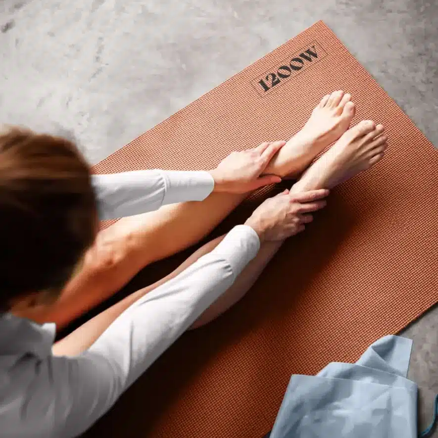 Person stretching on a brown yoga mat with 1200W branding in an indoor setting