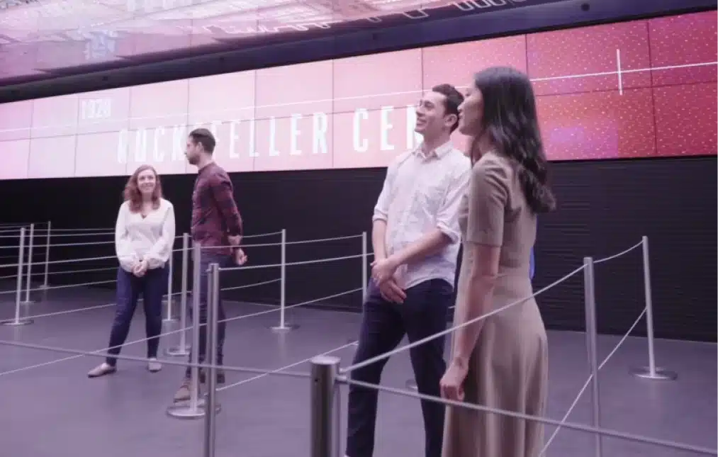 Visitors standing in line at Rockefeller Center with a digital timeline displayed overhead