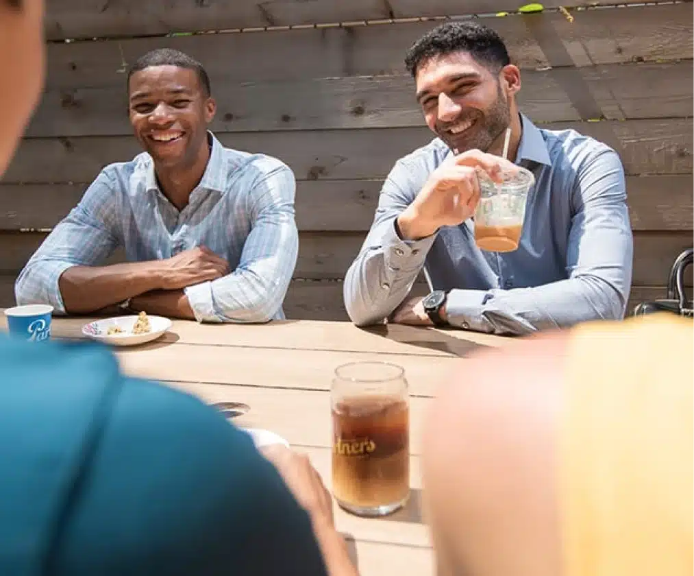 Two men laughing while enjoying iced coffee at an outdoor café table