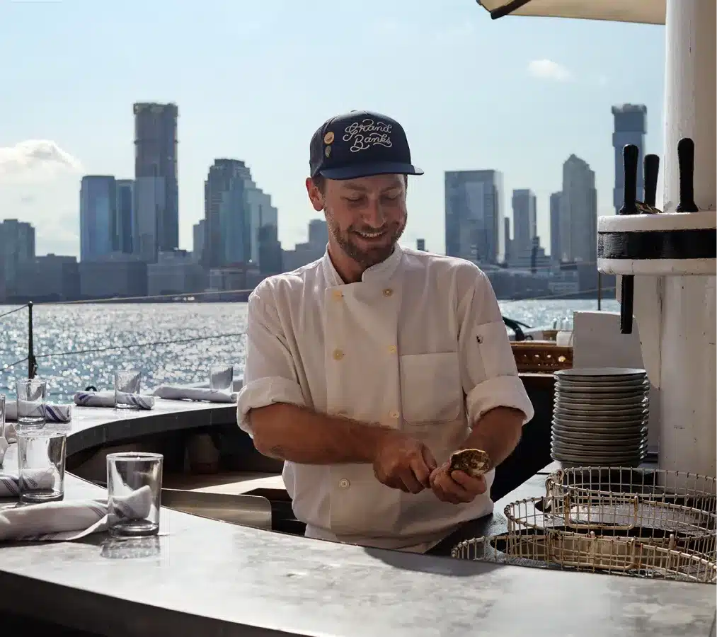 Smiling chef in a white uniform shucking oysters at an outdoor waterfront bar