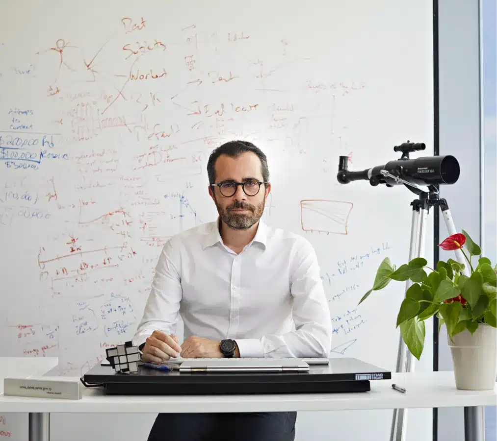 Businessman in glasses sitting at a desk with a whiteboard filled with handwritten notes