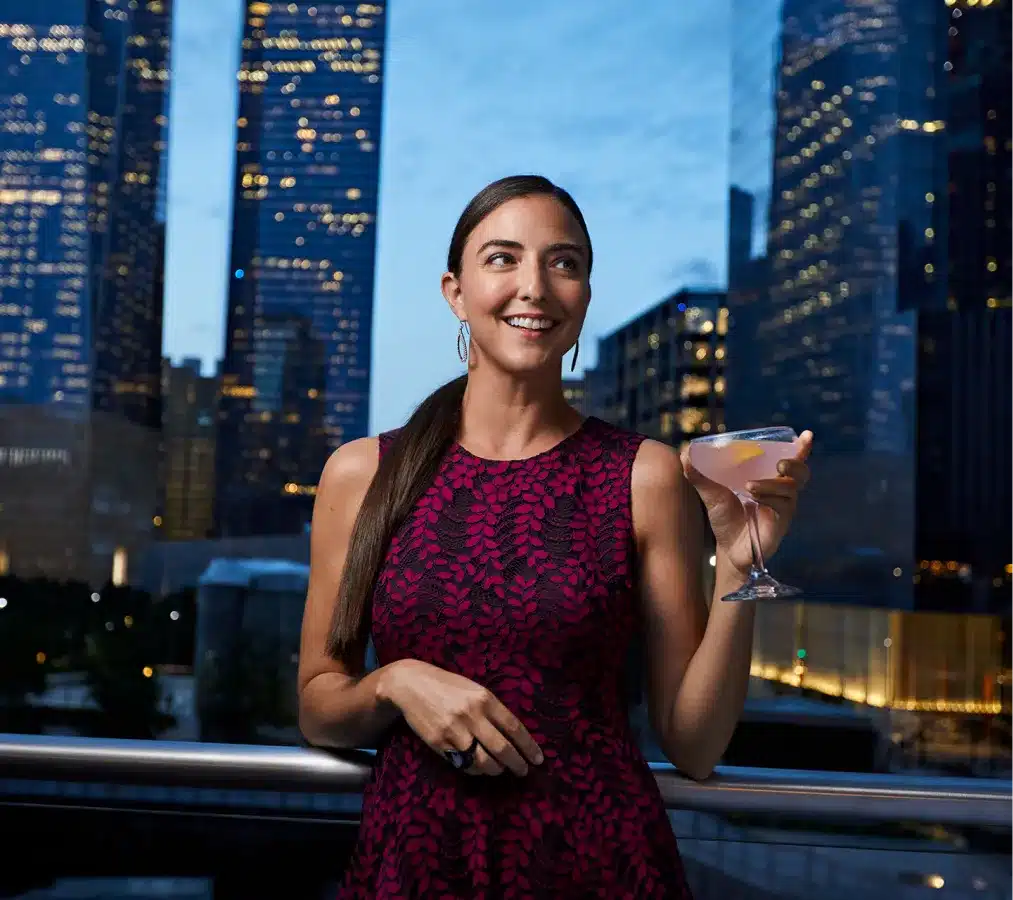 Woman in a magenta dress enjoying a cocktail on a rooftop with city skyscrapers at night
