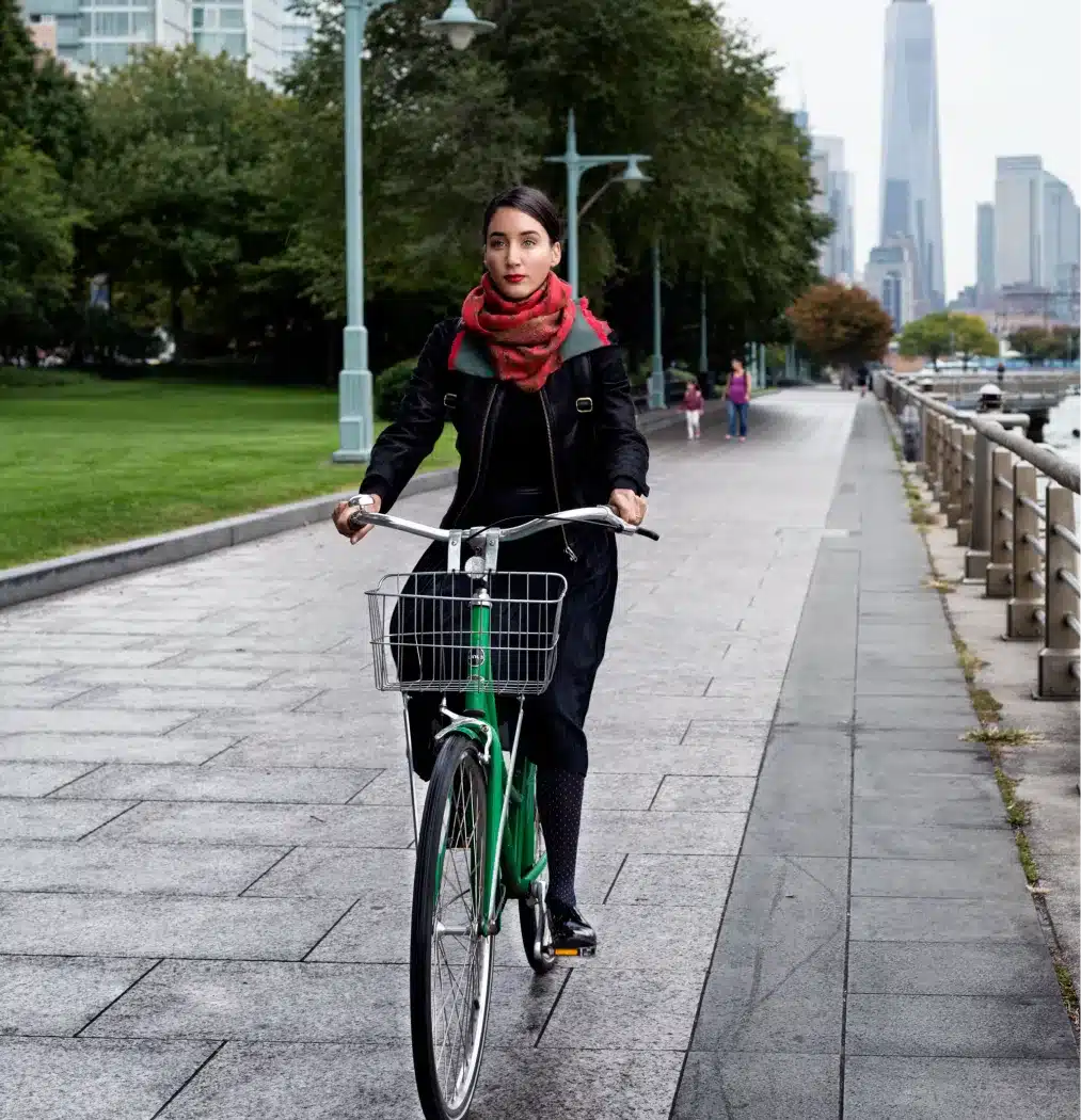 Woman riding a green bicycle along a waterfront pathway with a city skyline in the background