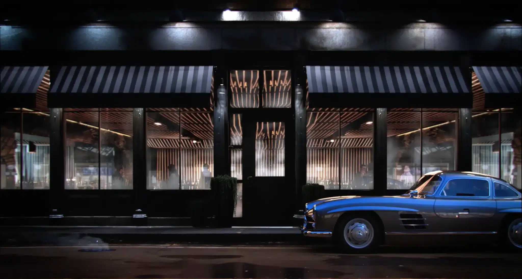 A classic blue Mercedes-Benz 300SL parked outside a stylish restaurant with glowing interiors