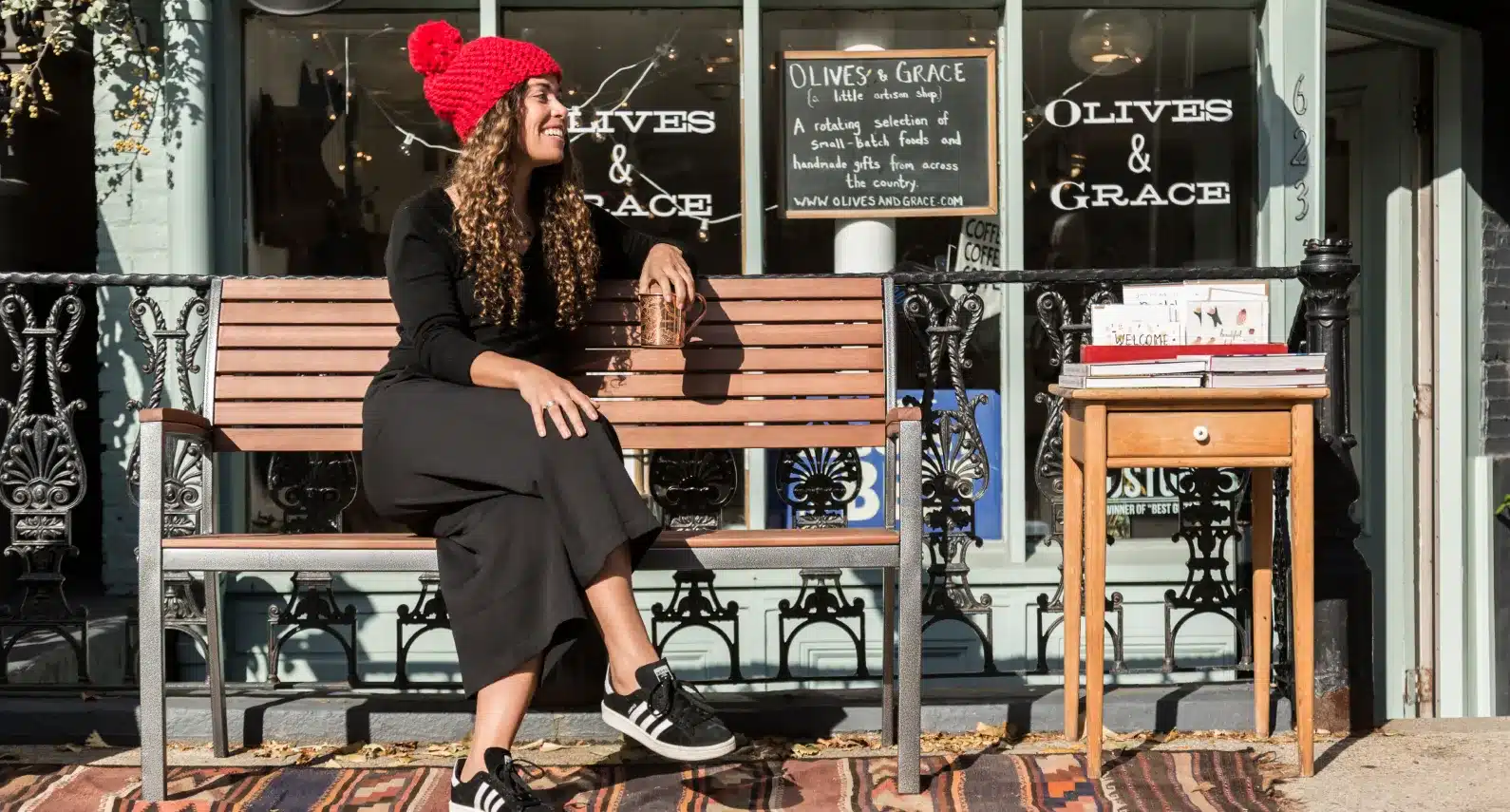 Smiling woman in a red beanie enjoying coffee outside Olives & Grace boutique