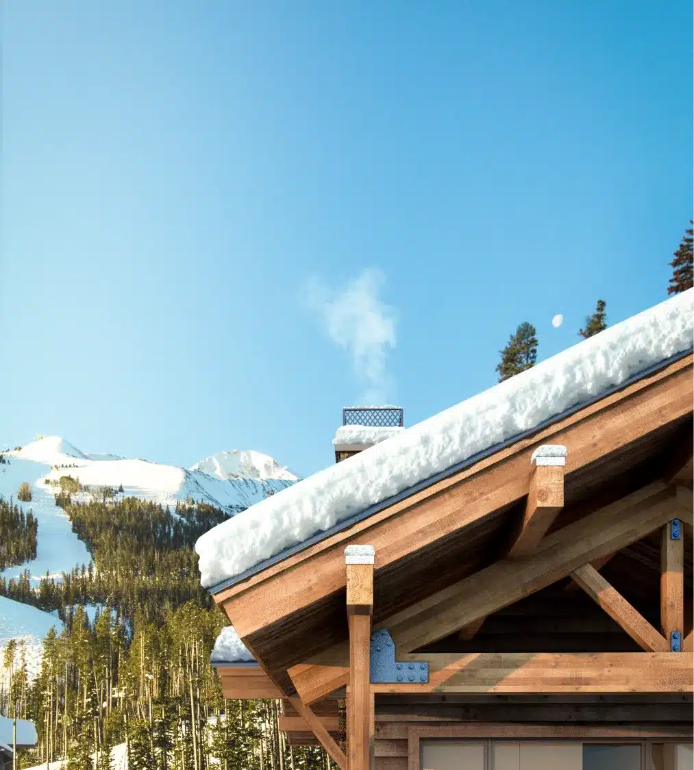 Snowy wooden chalet rooftop with chimney smoke rising against a clear blue mountain sky