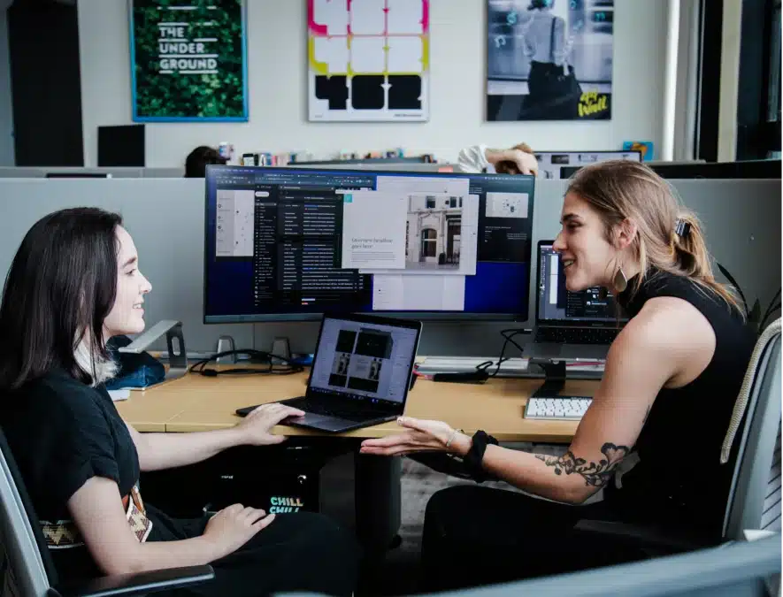 Office teammates collaborating over design files with dual monitors and open laptops in tech environment