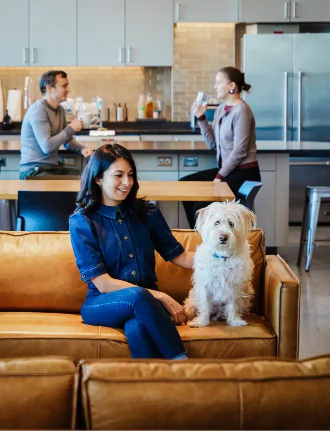 Woman relaxing on couch with friendly white dog while coworkers talk in modern kitchen background