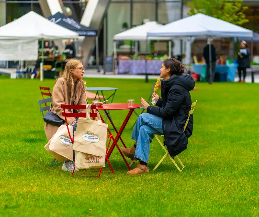 Two women with branded tote bags sitting at red metal table chatting during farmers market event