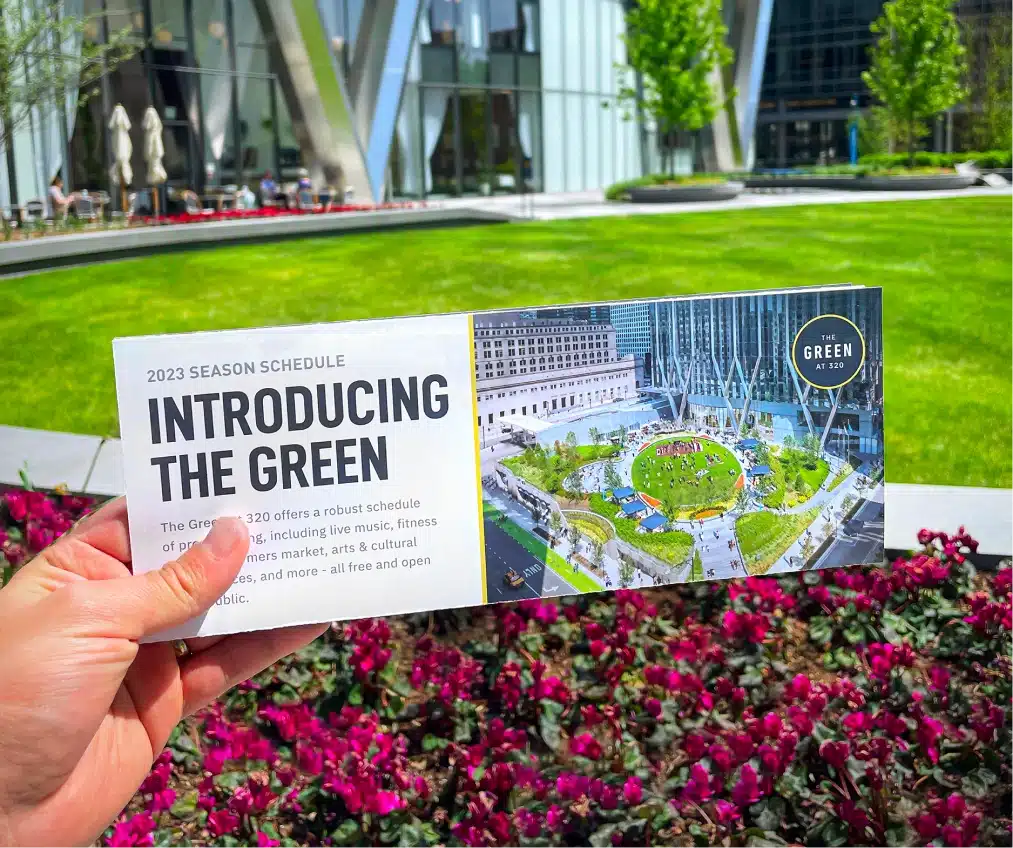 Hand holding brochure promoting The Green at 320, with blooming flowers and lawn in background