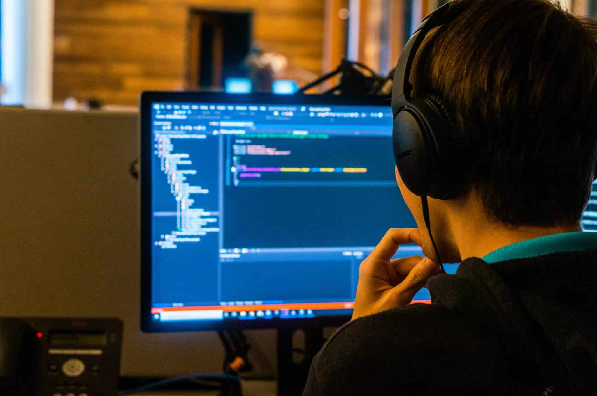 Developer wearing headphones working on code displayed on computer screen in dim office setting