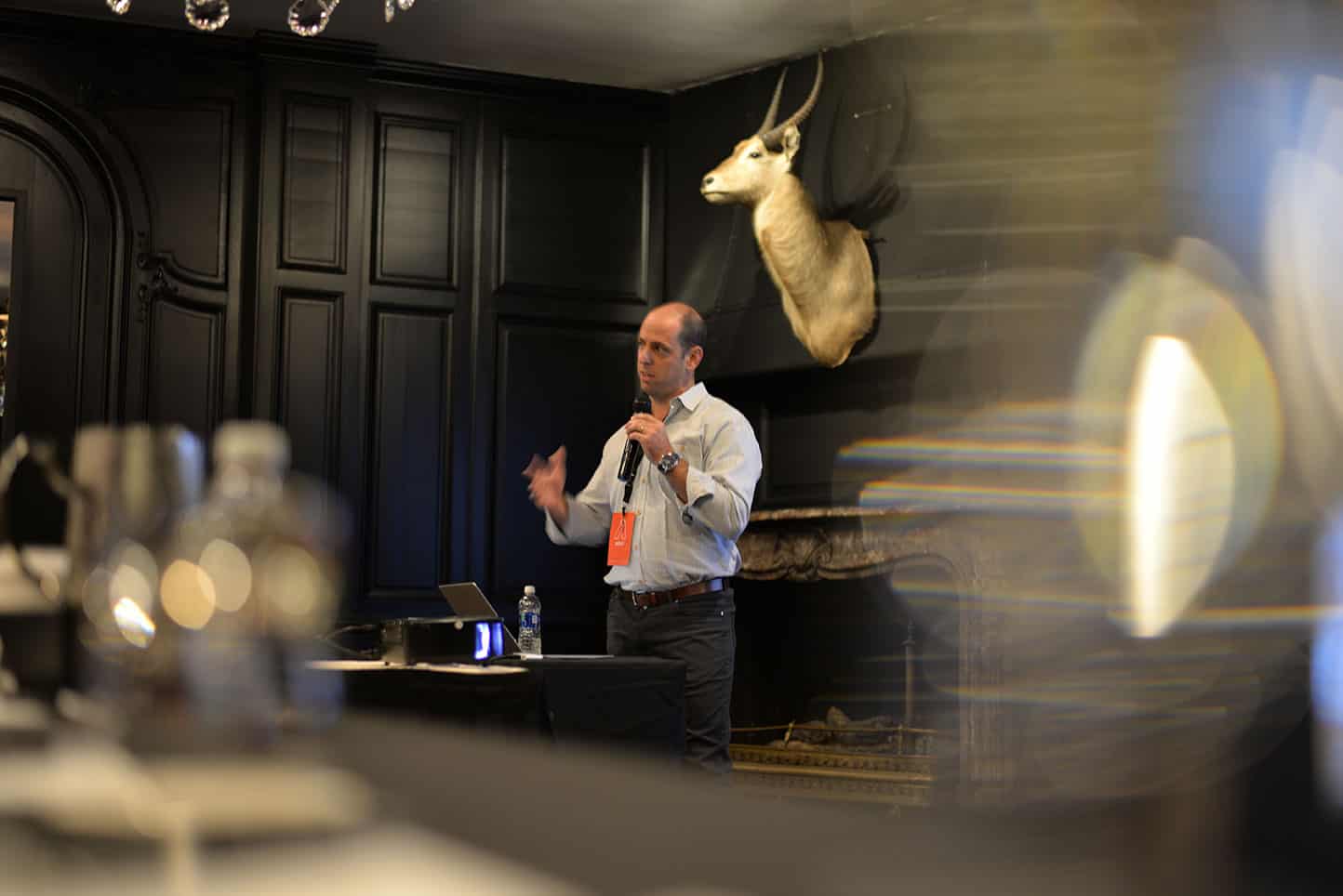 Man presenting at conference with microphone in moody black-paneled room and mounted antelope head
