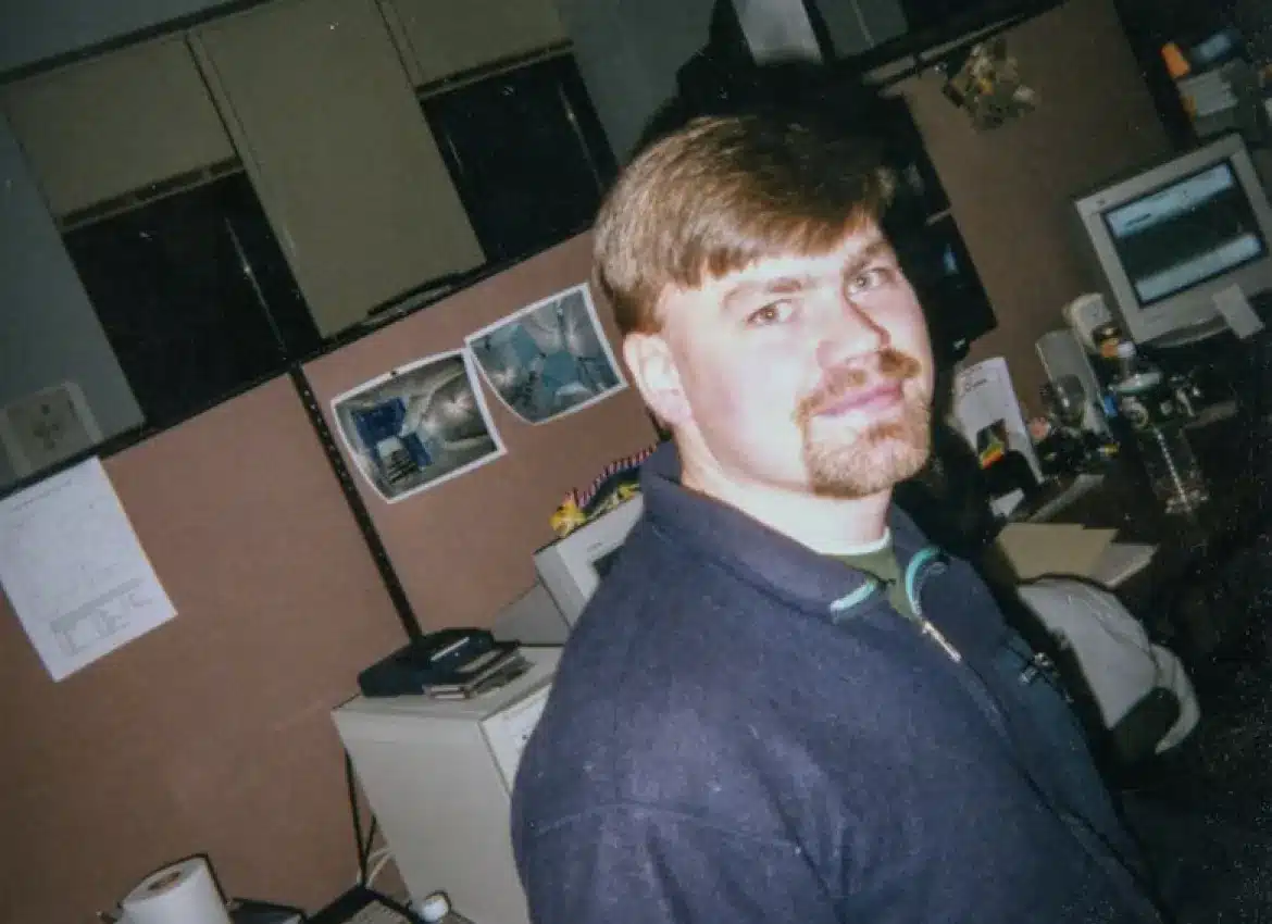 Young man with goatee standing near cubicle workspace, old CRT monitor in background