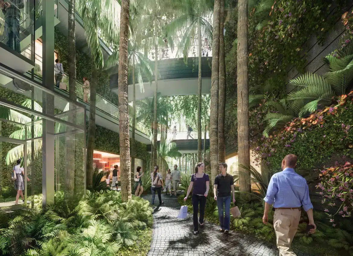Indoor garden walkway with tall trees, lush greenery, and glass architecture surrounding visitors