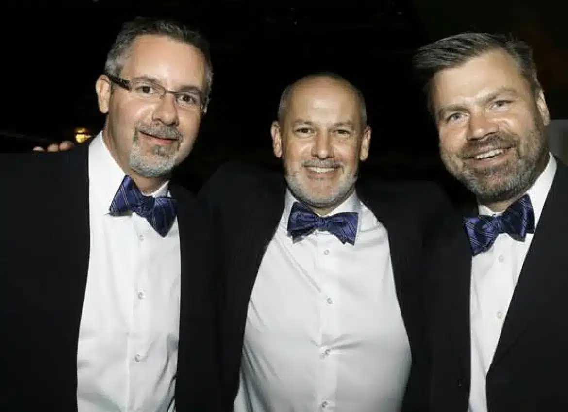 Three men in formal attire with bow ties posing together at elegant event