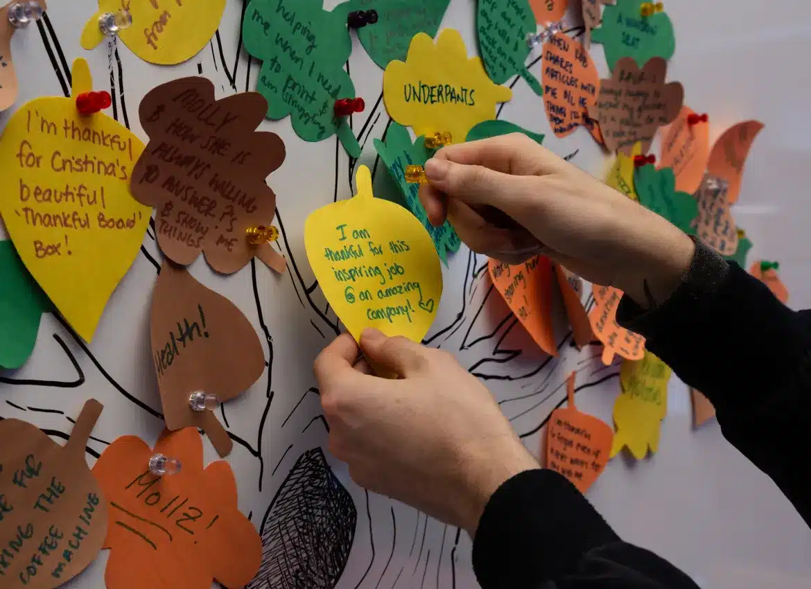 Hands pinning handwritten gratitude notes shaped like leaves on decorated wall board