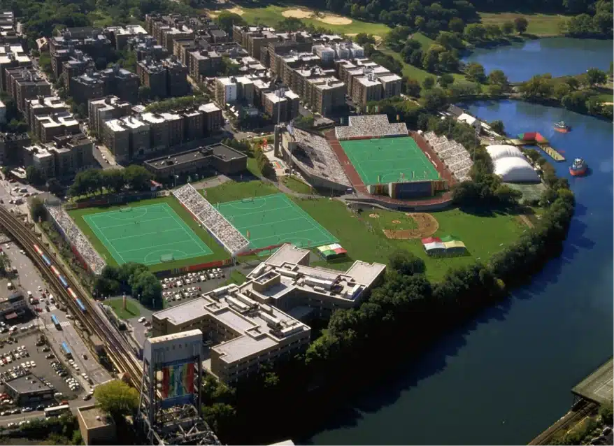 Aerial view of urban sports complex with multiple green fields near river and city buildings